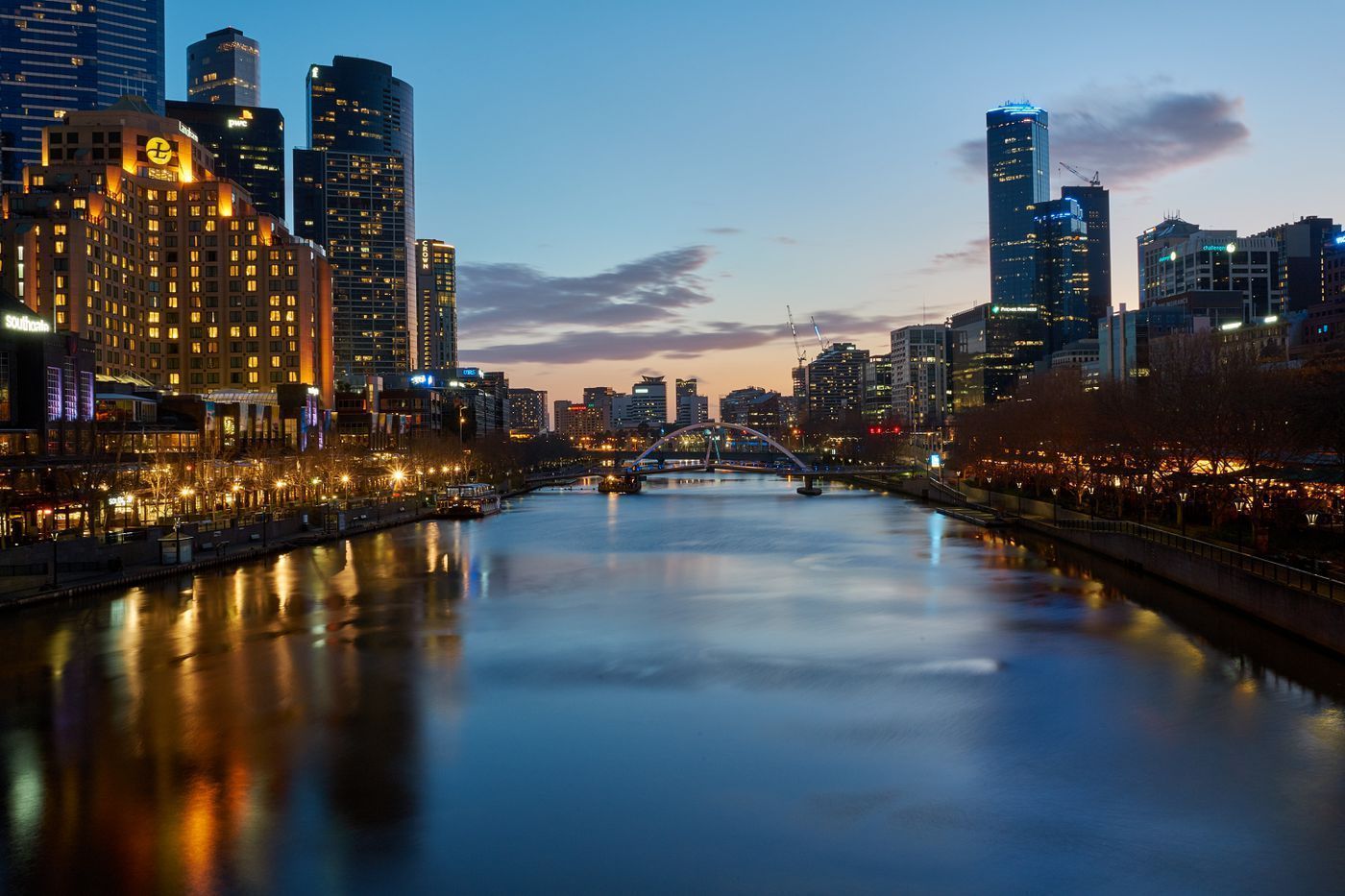 City skyline reflected in a river at dusk; buildings with illuminated windows line the riverbanks, dark blue water.