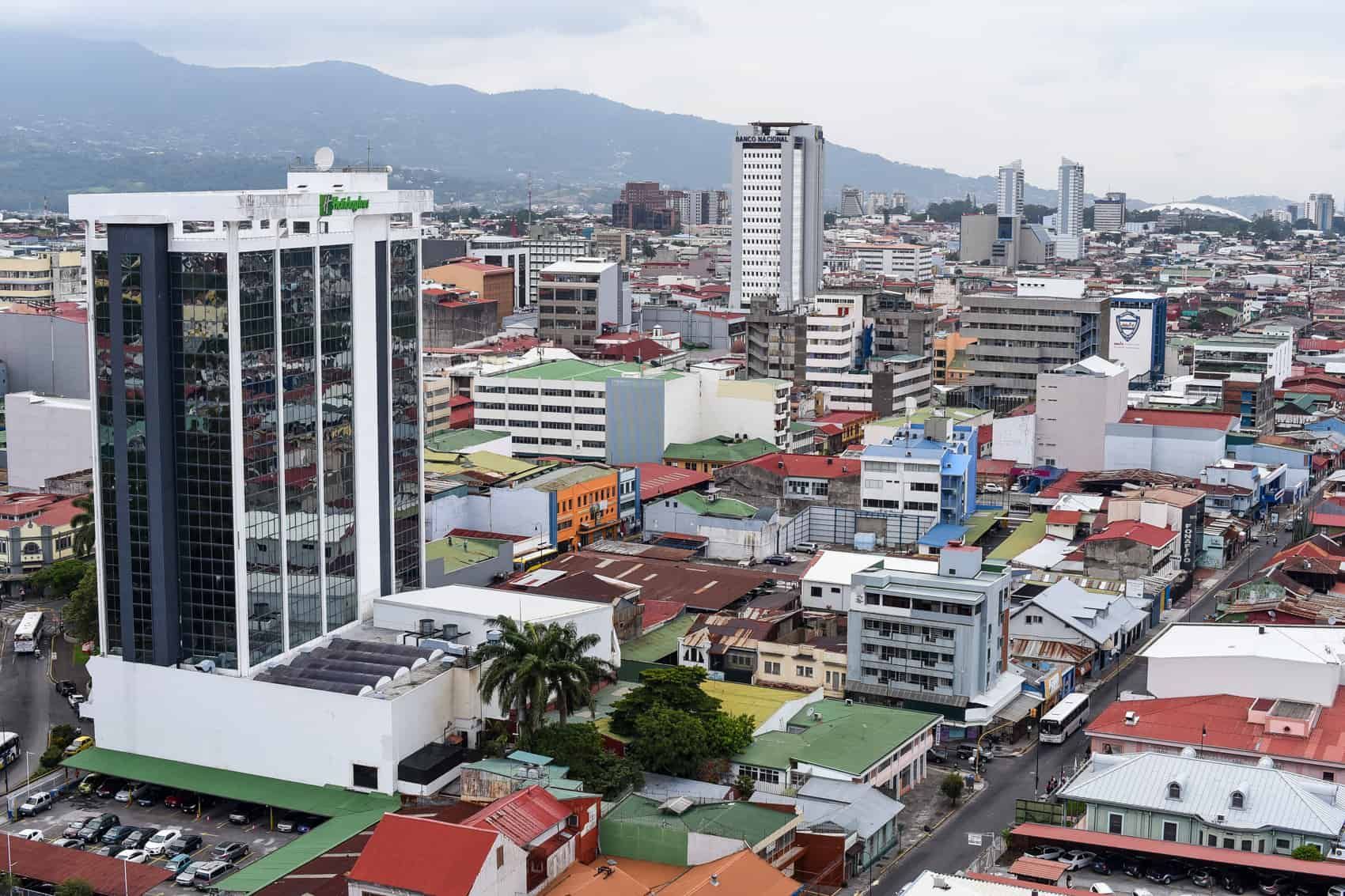 Cityscape of San José, Costa Rica, with tall buildings, colorful rooftops, and mountains in the background under an overcast sky.