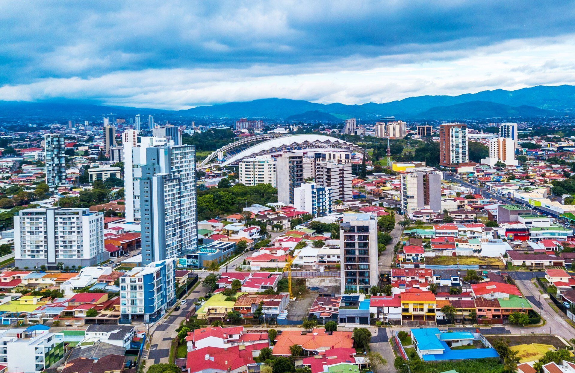Aerial view of San José, Costa Rica, with modern high-rise buildings, residential areas, a stadium, and mountains under a cloudy sky.