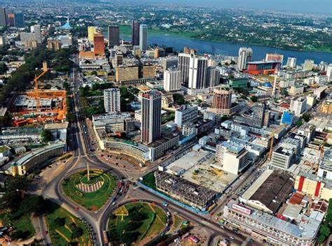 Aerial view of Abidjan, Côte d'Ivoire, featuring a cityscape of tall buildings, roads, roundabouts, and a river in the background.