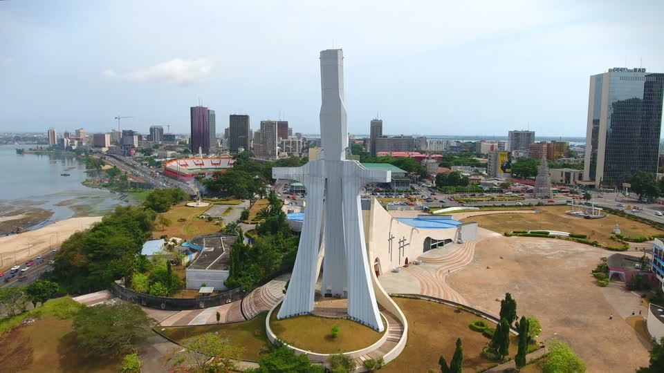 Aerial view of a white monument in Abidjan, Ivory Coast, with the city skyline and lagoon in the background under a cloudy sky.