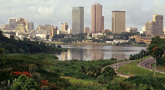 Abidjan skyline with tall buildings reflected in the lagoon. Green vegetation frames the view with a winding road in the foreground.
