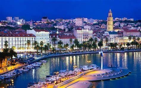 Night view of Split, Croatia, harbor with illuminated buildings, palm trees, and boats under a deep blue sky. A tall bell tower stands out.