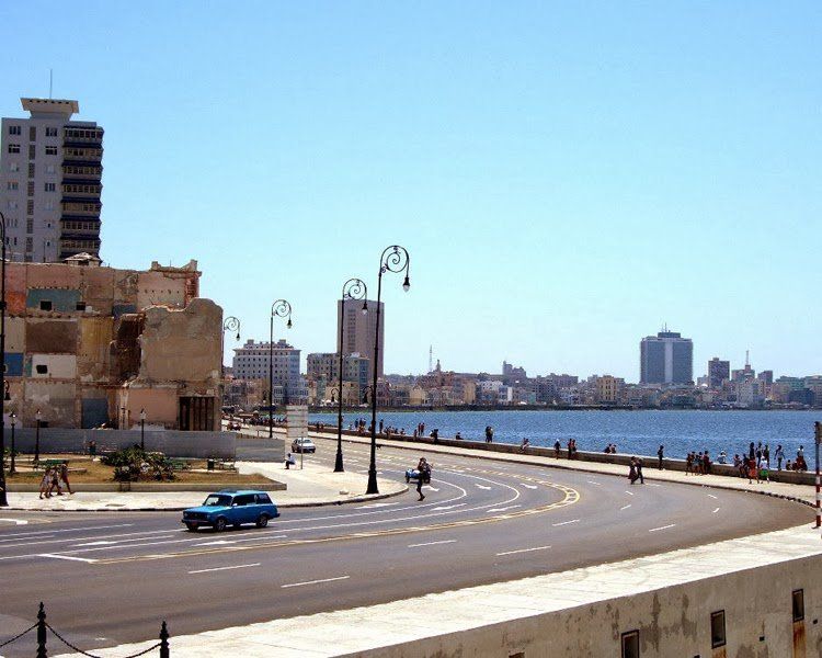 A coastal road in Havana, Cuba, with an ocean view, classic car, and buildings under a blue sky.