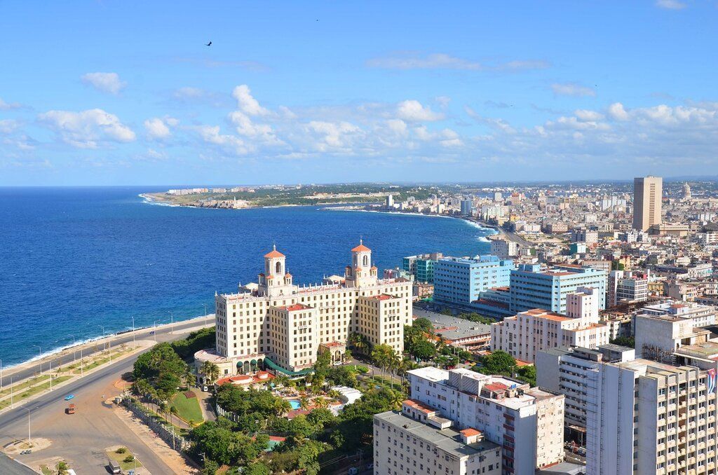 An aerial view of Havana, Cuba, with the ocean on the left, a large white building in the center, and city buildings filling the right side.