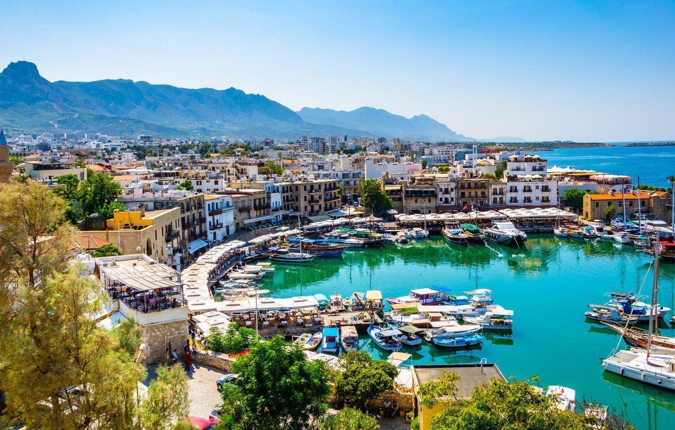 A coastal town with a harbor filled with boats, turquoise water, and buildings along the shore. Mountains are in the background.
