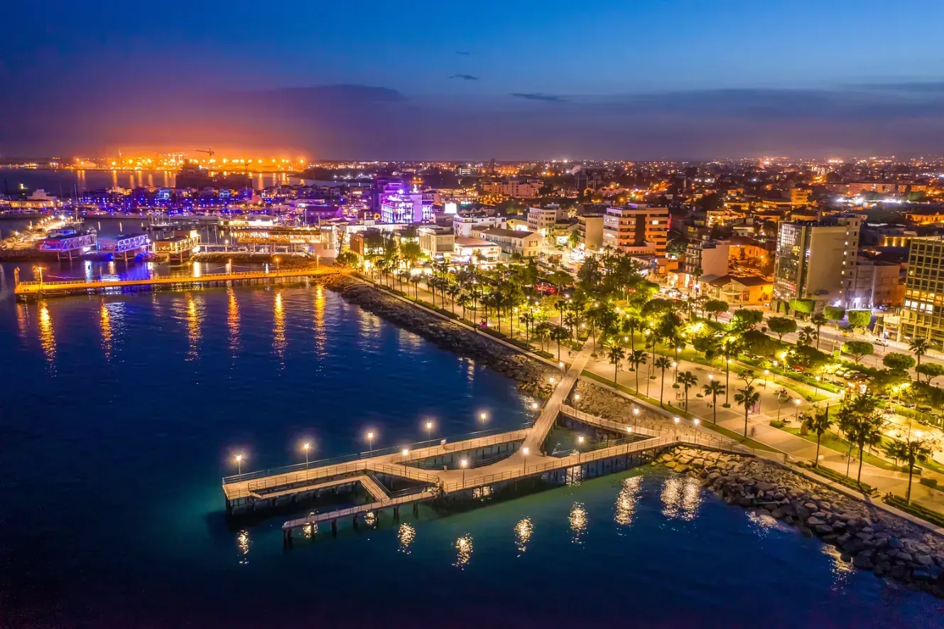 Night view of a city harbor with illuminated piers and buildings, along a coastline with palm trees, reflecting lights on dark blue water.