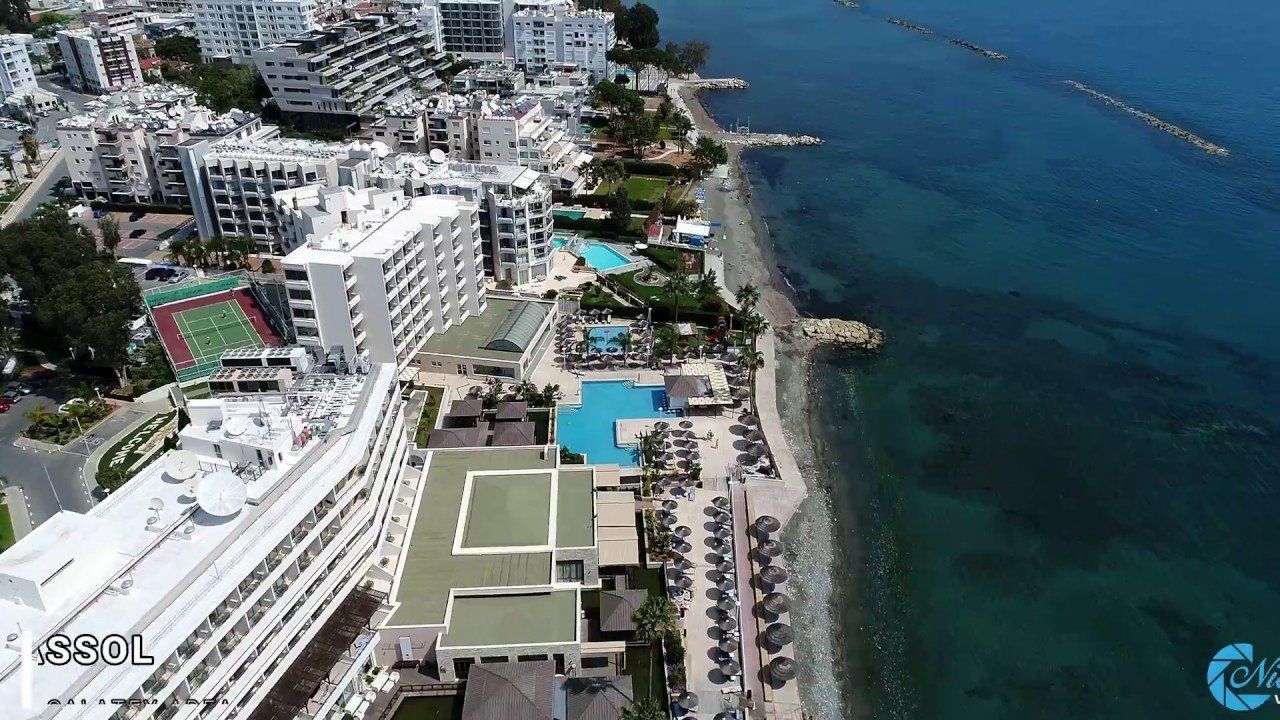 Aerial view of a white hotel complex next to a turquoise sea, with a swimming pool and beach.