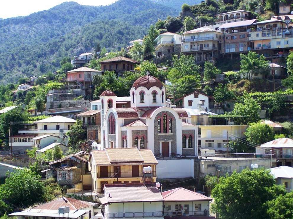 A church with white and red domes sits in a hillside village with houses, trees, and a mountain backdrop.