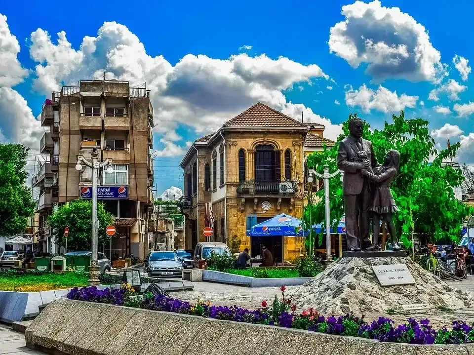 Statue of a man and child in a city square, with buildings, cars, and a blue sky with clouds.