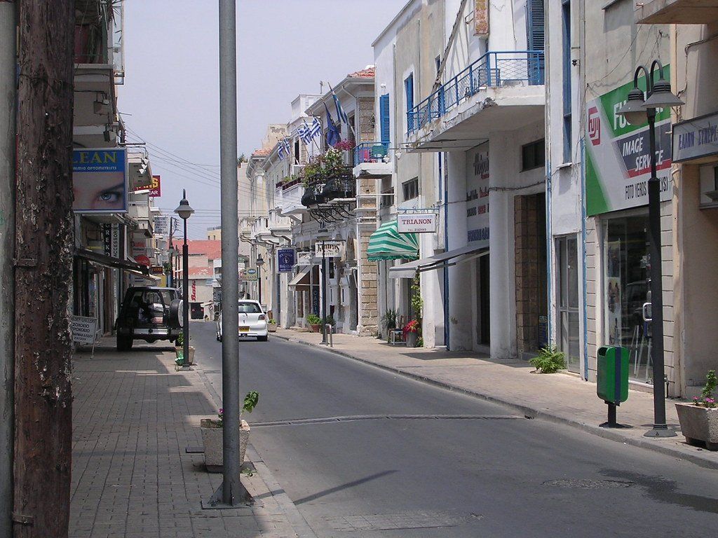A quiet street in a town with shops and balconies. Buildings are white and beige, with blue accents. A car drives down the center.