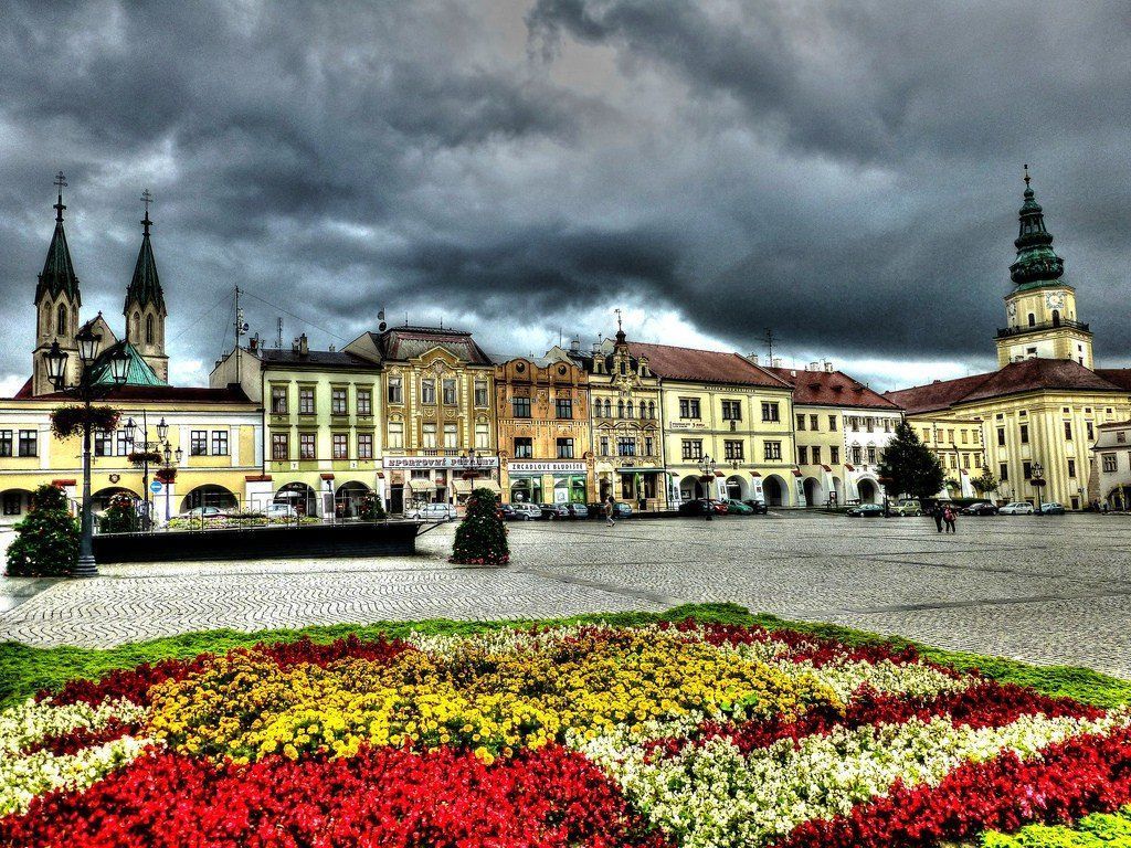 A colorful flower bed in front of a European town square with ornate buildings under a dramatic, cloudy sky.