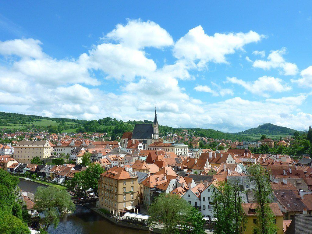 View of Cesky Krumlov, Czech Republic, with red-roofed buildings, a church steeple, and a river under a blue sky with puffy clouds.