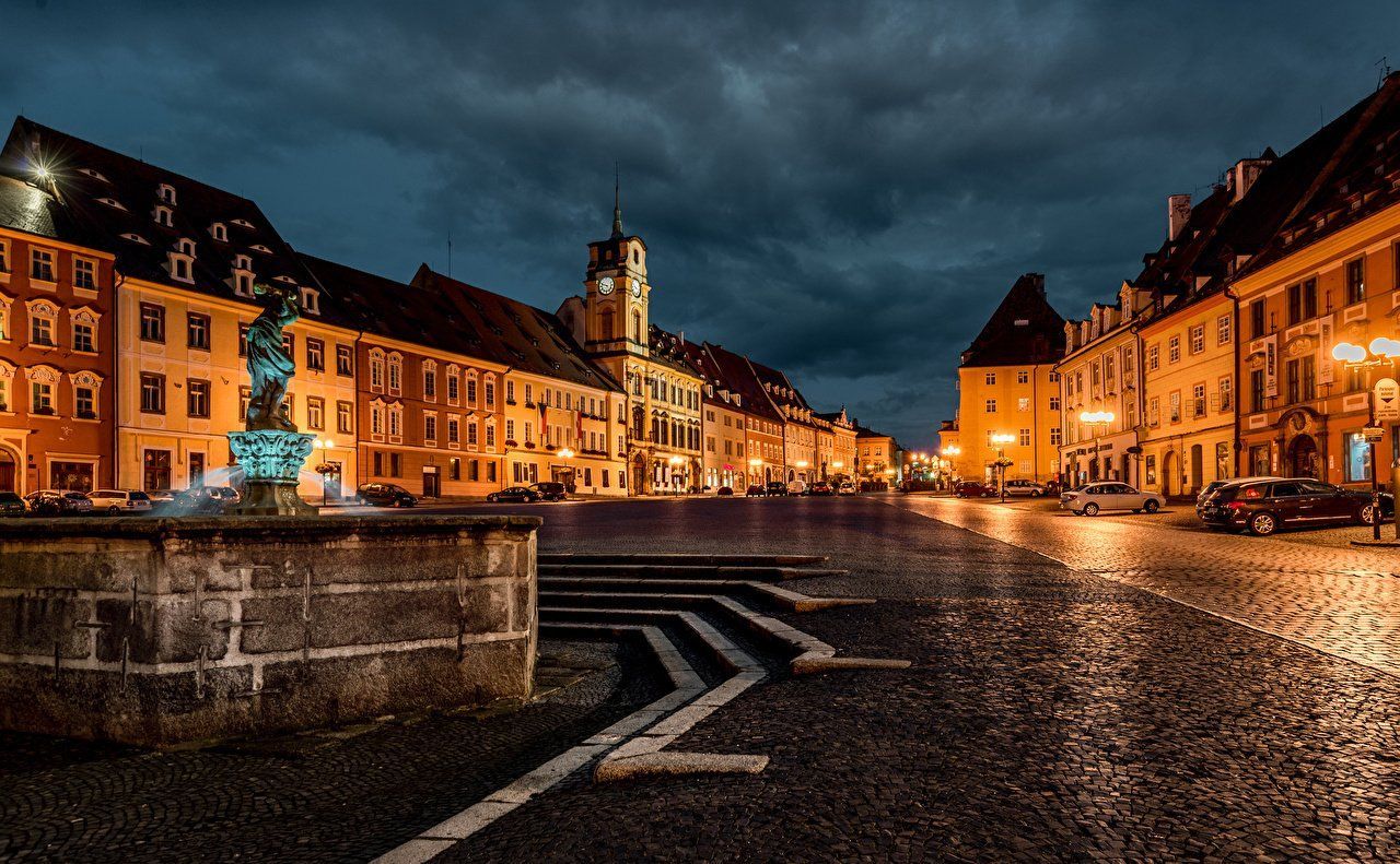 Cobblestone square at dusk, lit by streetlights. Buildings line the square; a central fountain sits in the foreground under a cloudy sky.