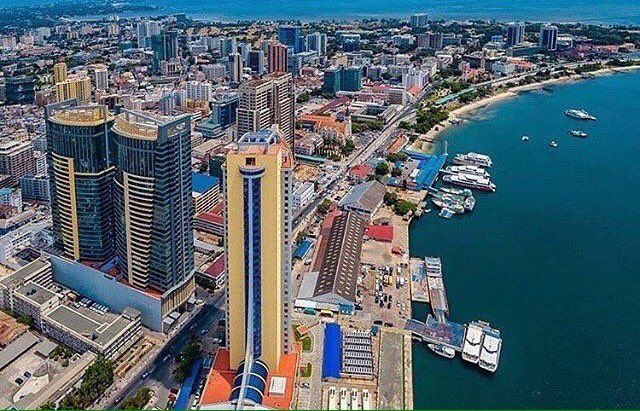 Aerial view of Dar es Salaam, Tanzania, with skyscrapers, a harbor filled with boats, and a coastal cityscape under a bright sky.