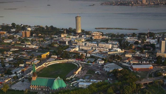 Aerial view of Pelotas, Brazil, showing a church with a green roof, a racetrack, and a tall water tower near a bay with city buildings in the distance.