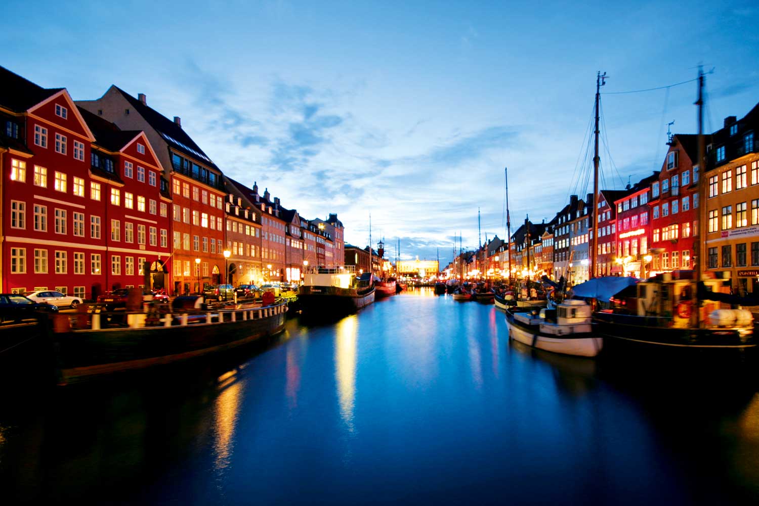 Canal lined with colorful buildings, lit at dusk. Boats are docked along the water, reflecting the lights.