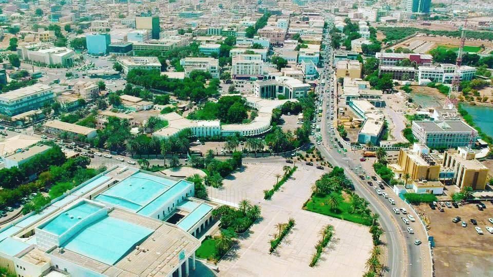 Aerial view of a city with a wide boulevard lined with cars, buildings, and greenery. Brightly lit, mostly neutral colors.