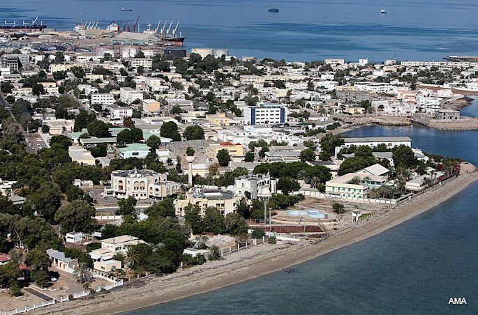 Aerial view of a coastal city with buildings, trees, and a harbor, likely in a warm climate. Blue water surrounds the land.