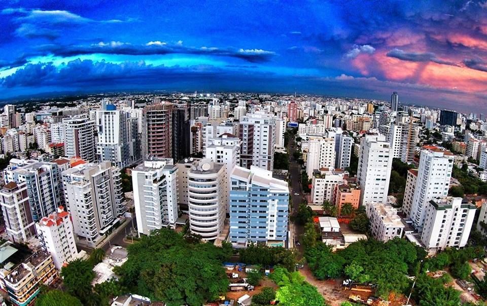 Aerial view of a dense city with tall buildings and green trees under a dramatic, colorful sky.
