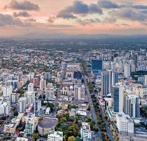 Aerial view of Santo Domingo, Dominican Republic, showcasing a modern cityscape with tall buildings and a central road lined with trees under a sunset sky.