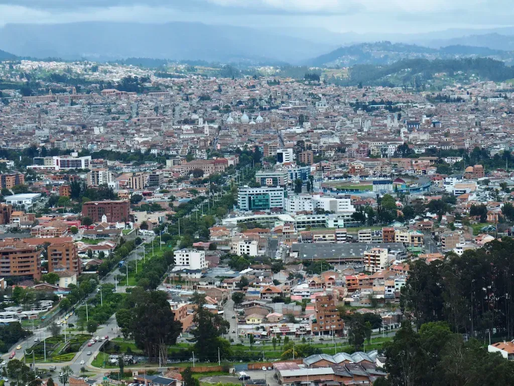 An aerial view of a sprawling city with buildings, roads, and green spaces, set against a backdrop of mountains under a cloudy sky.