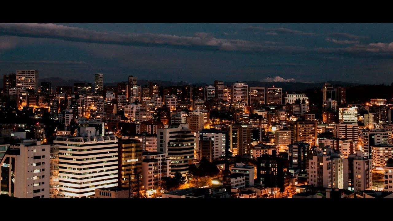 Nighttime cityscape with illuminated buildings under a dusky sky. Tall buildings and apartment blocks fill the frame, suggesting an urban environment.