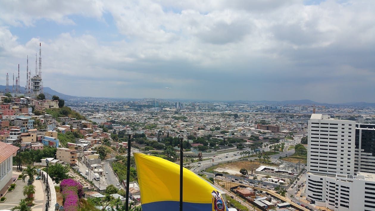 Cityscape under a cloudy sky, with a yellow and blue flag in the foreground. Buildings and communications towers are visible.