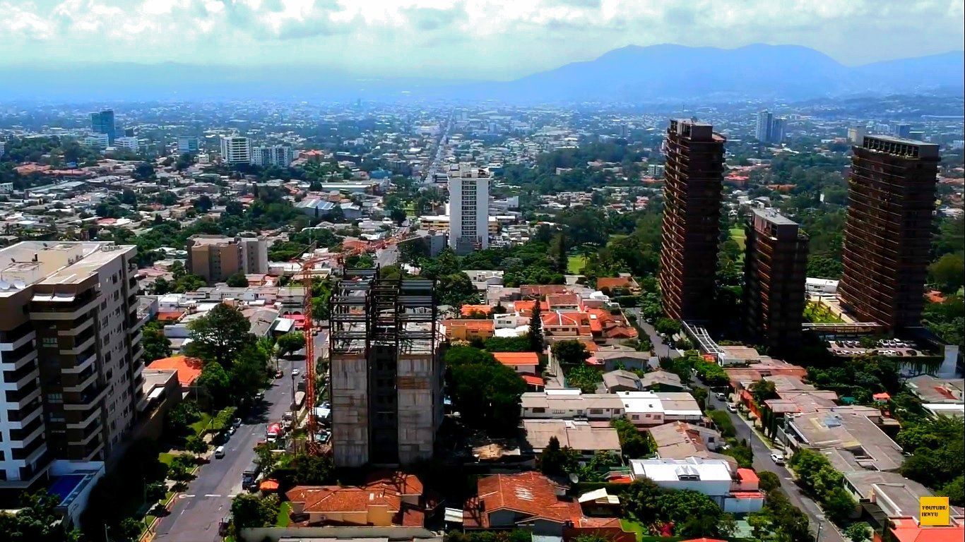 Aerial view of a dense city, with tall buildings and tree-lined streets, under a partly cloudy sky.