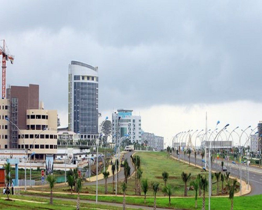 Modern cityscape with high-rise buildings, roads, and green spaces under a cloudy sky. A crane is visible.