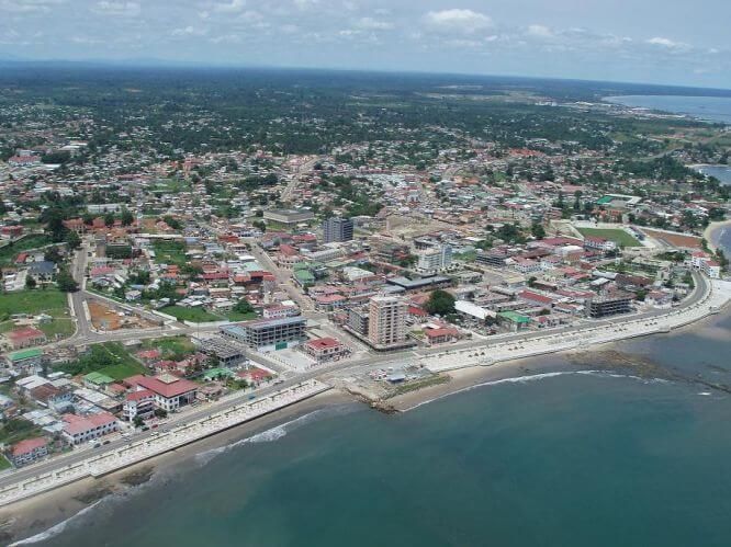 Aerial view of a coastal town with buildings of various colors, a wide road along the shore, and green ocean.