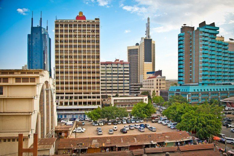 Skyline of Nairobi, Kenya, featuring tall buildings of various colors, parked cars, and a vibrant cityscape under a bright sky.
