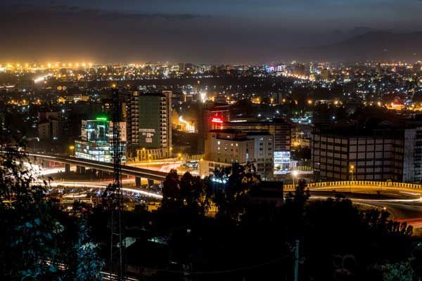 Nighttime cityscape with illuminated buildings and roads; orange and white lights dot the horizon.