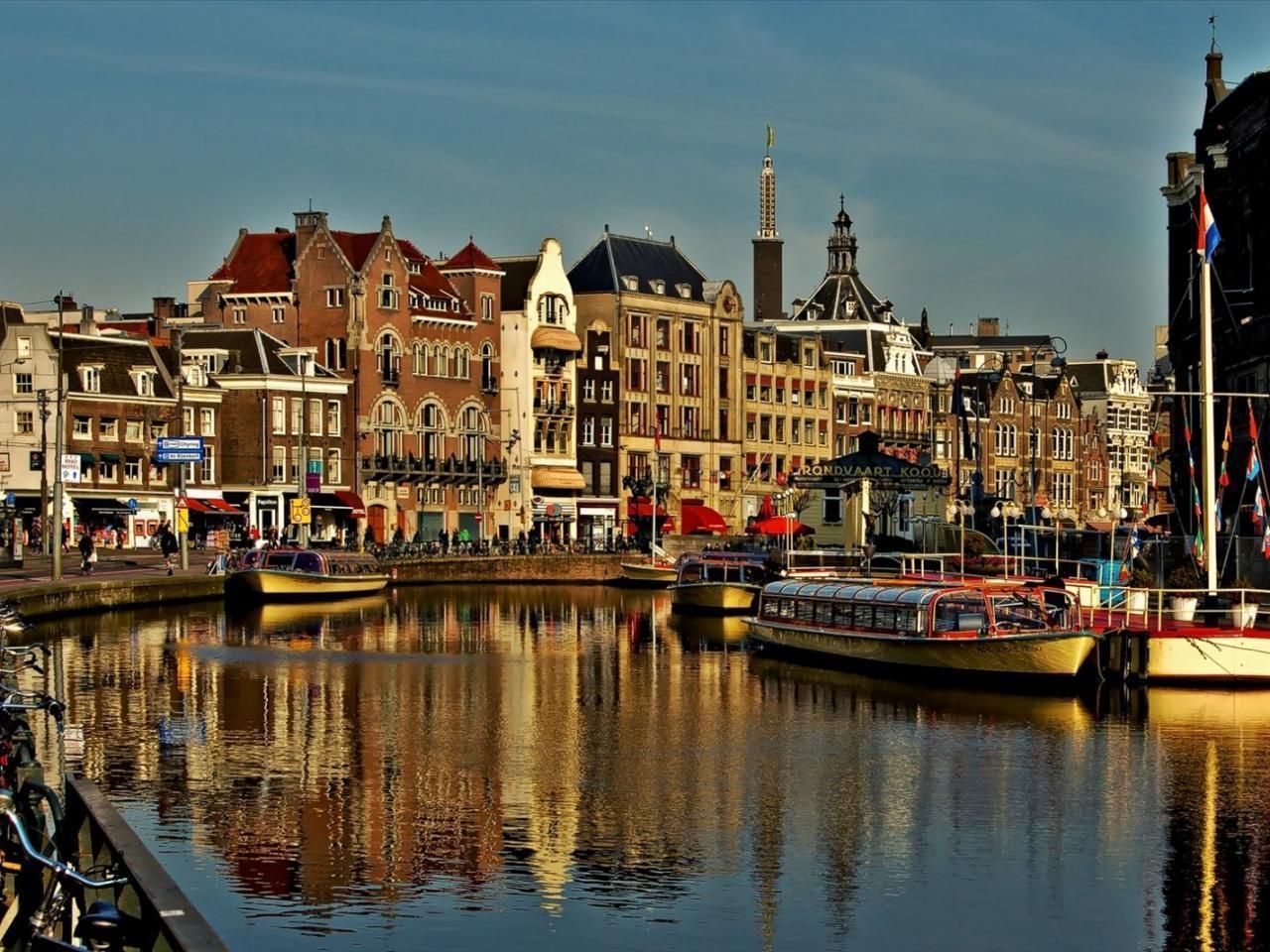 Canal in Amsterdam with colorful buildings reflecting in the water, boats docked along the banks, and a clear, sunny sky.