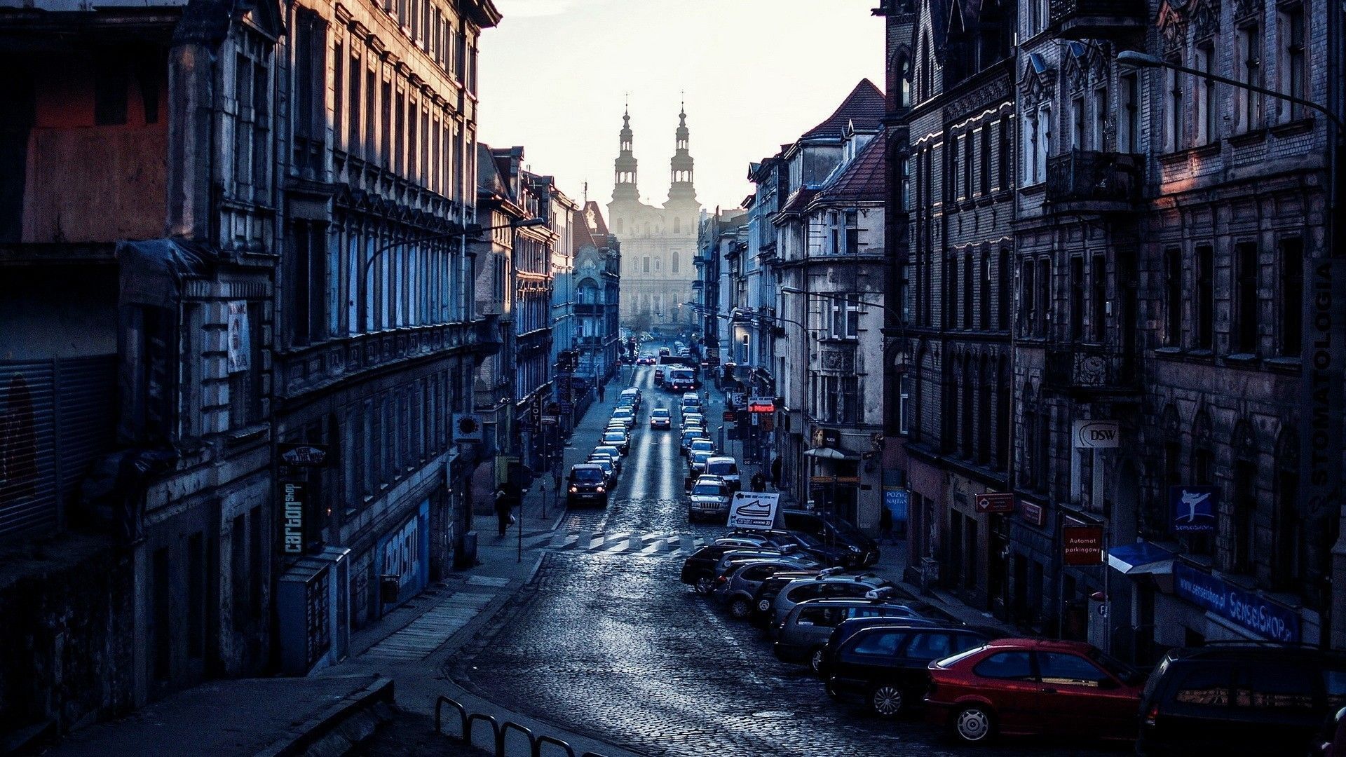 A European city street with old buildings, cars, and a cathedral in the distance; shot at dusk with a blue-gray color palette.