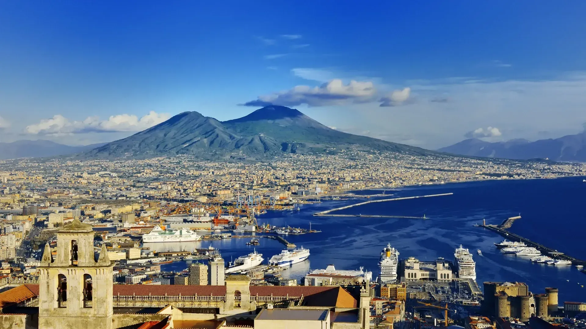 View of Naples, Italy with Mount Vesuvius in the background. Cityscape and harbor dominate the foreground under a blue sky.