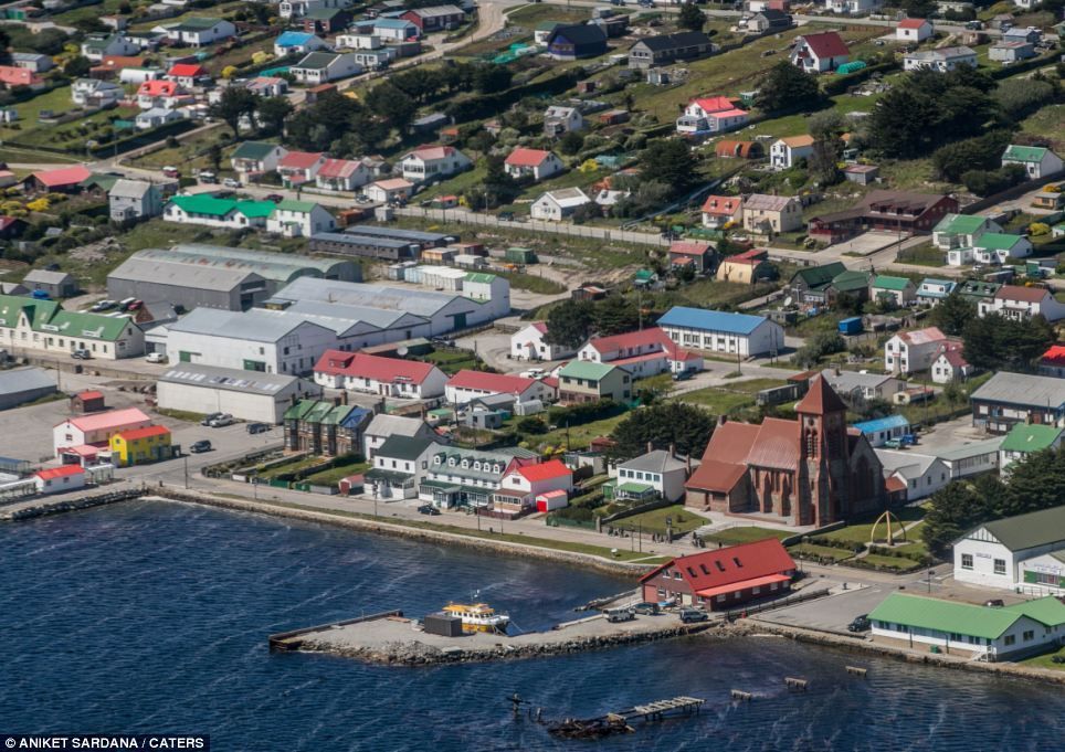 Aerial view of Stanley, Falkland Islands. Colorful buildings line the harbor and hillside, with a prominent red brick church and pier in the foreground.