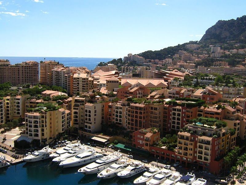 Marina filled with yachts, buildings lining the harbor in Monaco, with hillside residences in the background, blue sky.