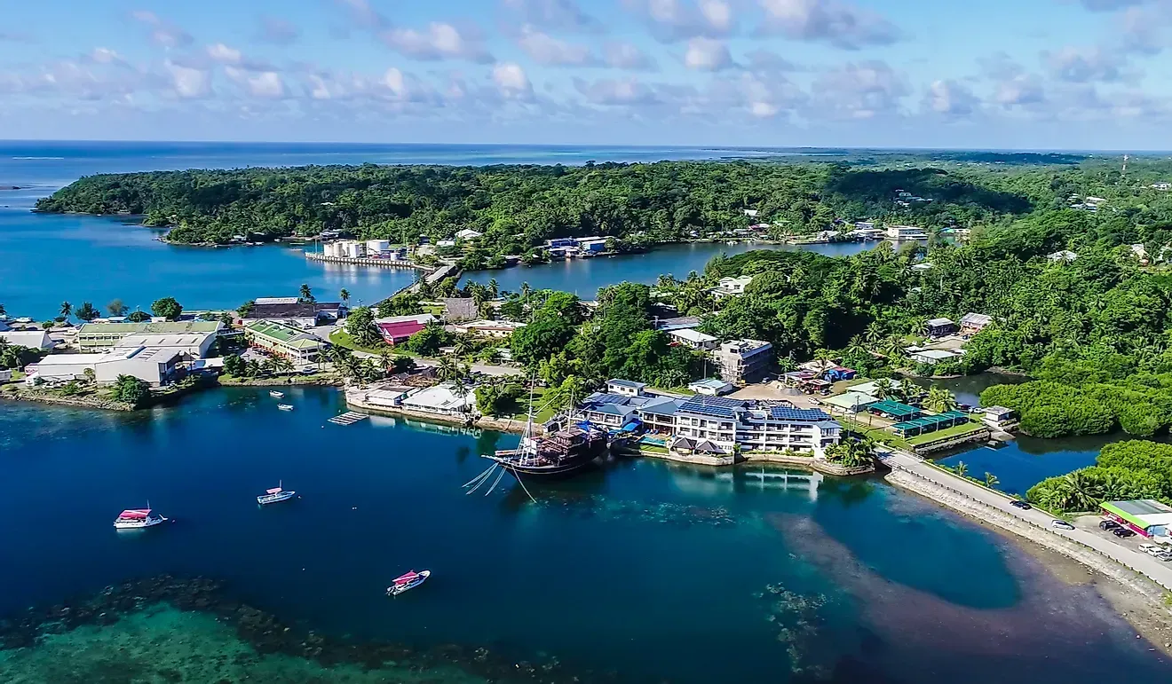 Aerial view of a coastal town with blue waters, lush green islands, and a bridge. Boats and buildings dot the shoreline.
