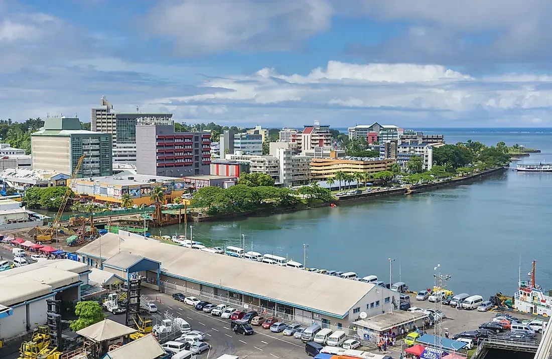 An aerial view of Apia, Samoa, with buildings along the waterfront, a dock, and a harbor on a sunny day.
