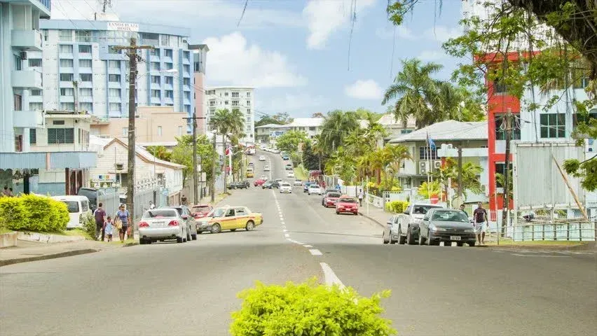Street scene in a town with cars, buildings of varying heights, and palm trees under a partly cloudy sky.