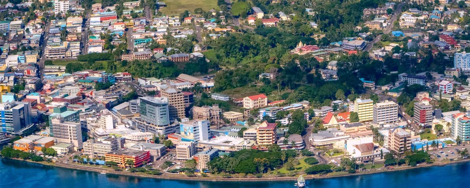 Aerial view of a coastal city with colorful buildings and lush green foliage. A body of blue water sits in the foreground.