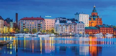 Waterfront cityscape at dusk with buildings, harbor, and reflections. Features buildings with red roofs and a large church with a dome.
