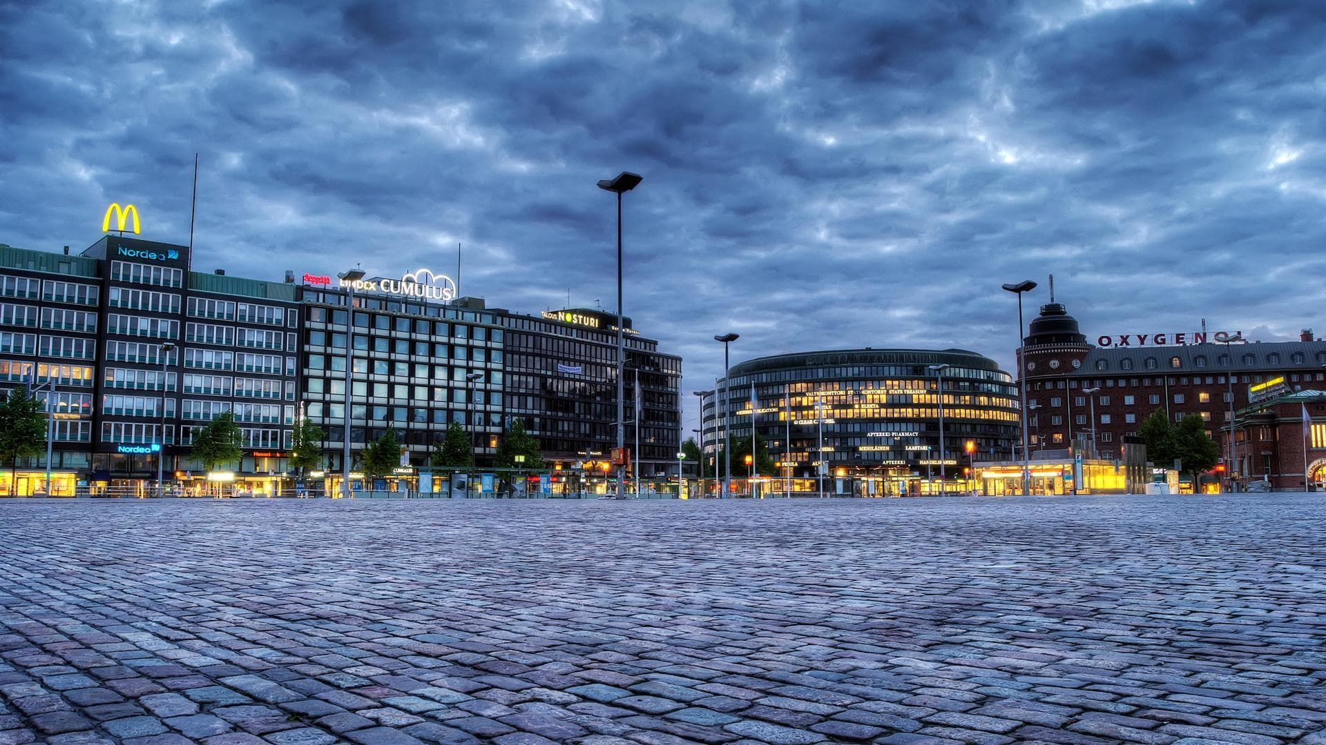 Cobblestone square in front of city buildings at dusk, under a cloudy sky. Features a McDonald's sign.