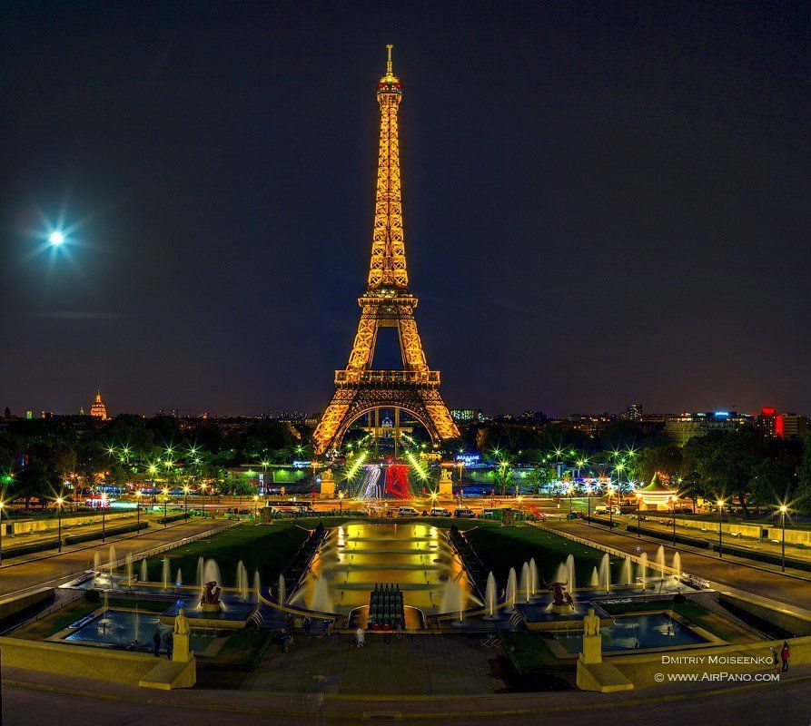 Eiffel Tower lit up at night, with a moon in the sky and fountains in the foreground.