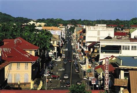 View down a street in Cayenne, French Guiana, lined with buildings of varying heights and colors, with cars and trees.