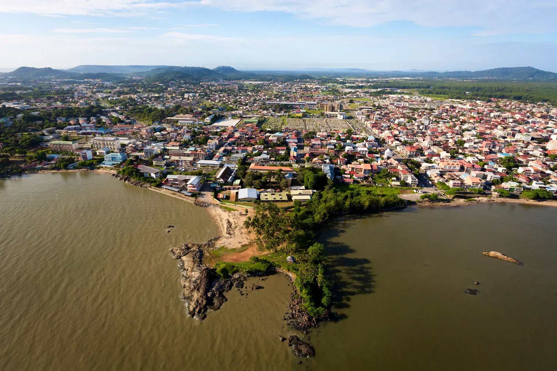 Aerial view of a coastal city with brown water, dense buildings, and green vegetation.