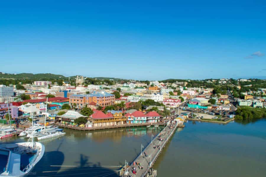 Aerial view of St. John's, Antigua. Waterfront promenade with colorful buildings and a bridge over the harbor under a bright blue sky.