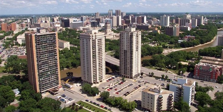 Aerial view of a city skyline with tall buildings, green trees, and a river running through it. The focus is on three high-rise apartment buildings in the foreground.