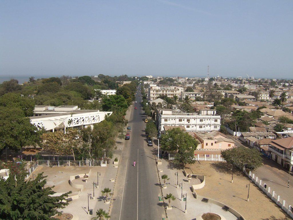 A wide, tree-lined avenue in Dakar, Senegal, stretches towards the ocean, flanked by buildings and rooftops under a clear sky.